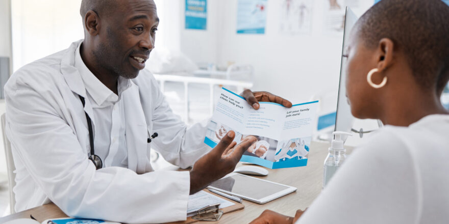 A doctor giving patient hospital information at a clinic and explaining medical benefits to a woman in his office. African American GP or healthcare professional having a discussion with a female.