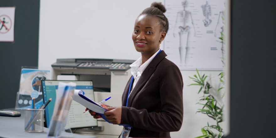 Portrait of female receptionist working at hospital reception counter with health insurance reports. Medical worker using computer to make appointments and fill in form papers at registration desk.